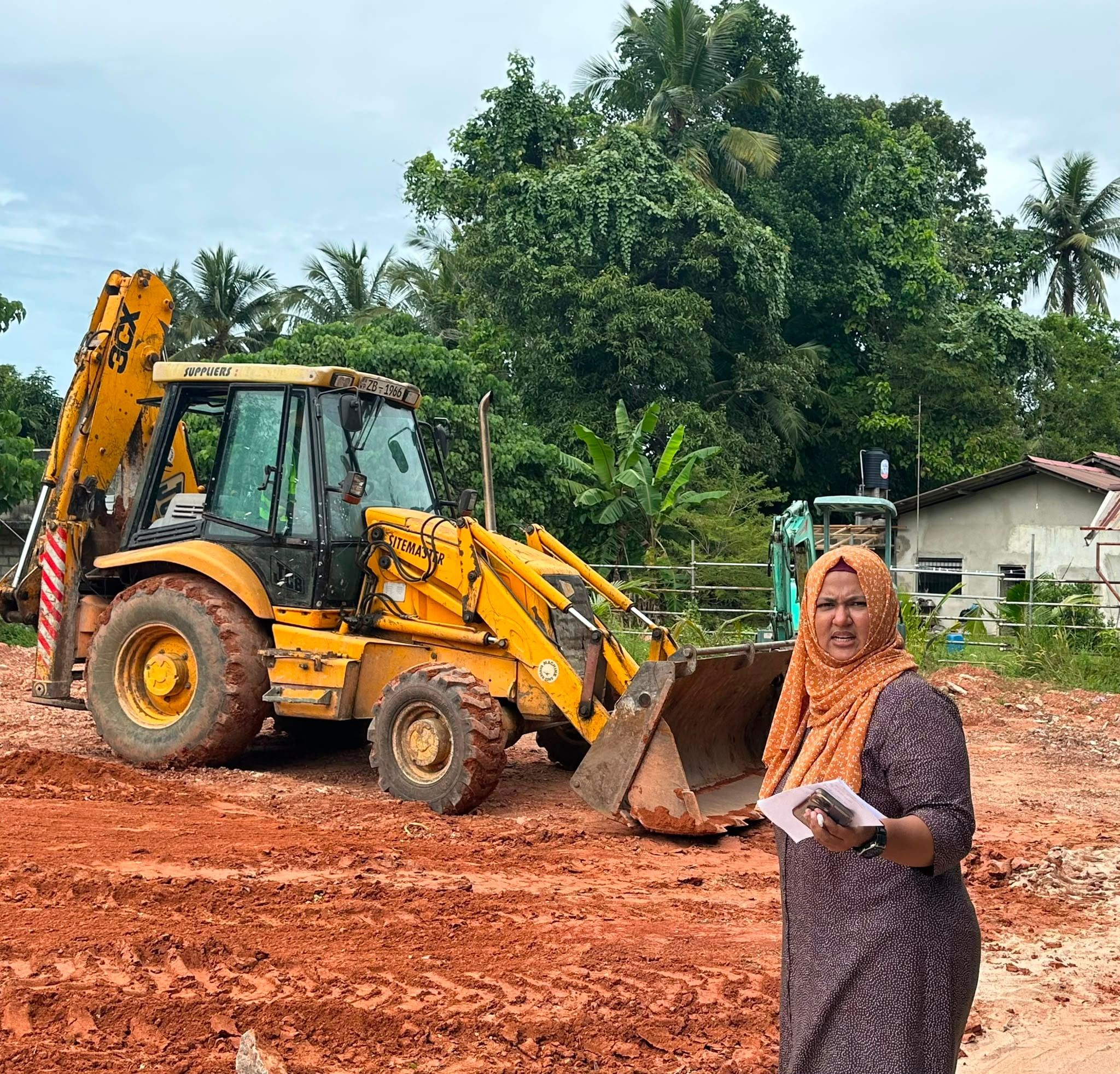 A yellow backhoe loader on cleared red earth under palm trees, a woman in an orange headscarf in front holding papers and a phone