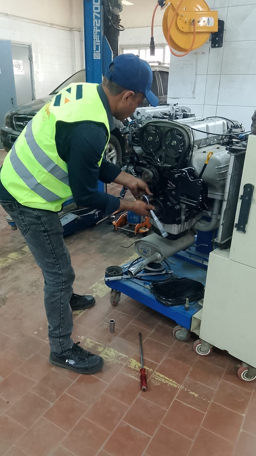 A mechanic in a Eurozone high-visibility vest and a cap bending to work on an engine mounted on a stand, tools on the tiled workshop floor