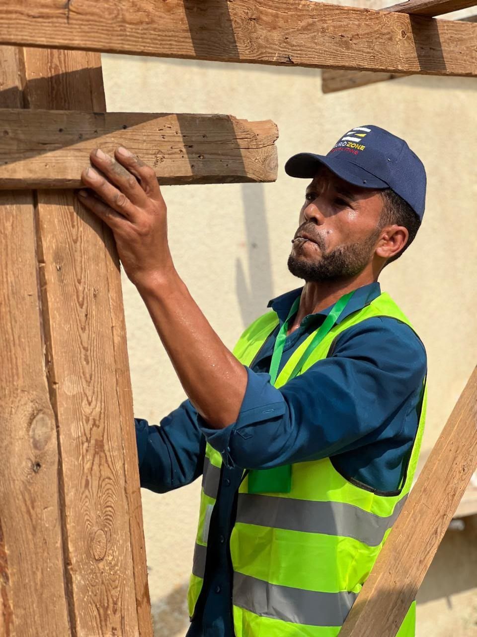 A worker in a Eurozone cap and hi-vis vest reaching up to fit a timber beam into a wooden frame