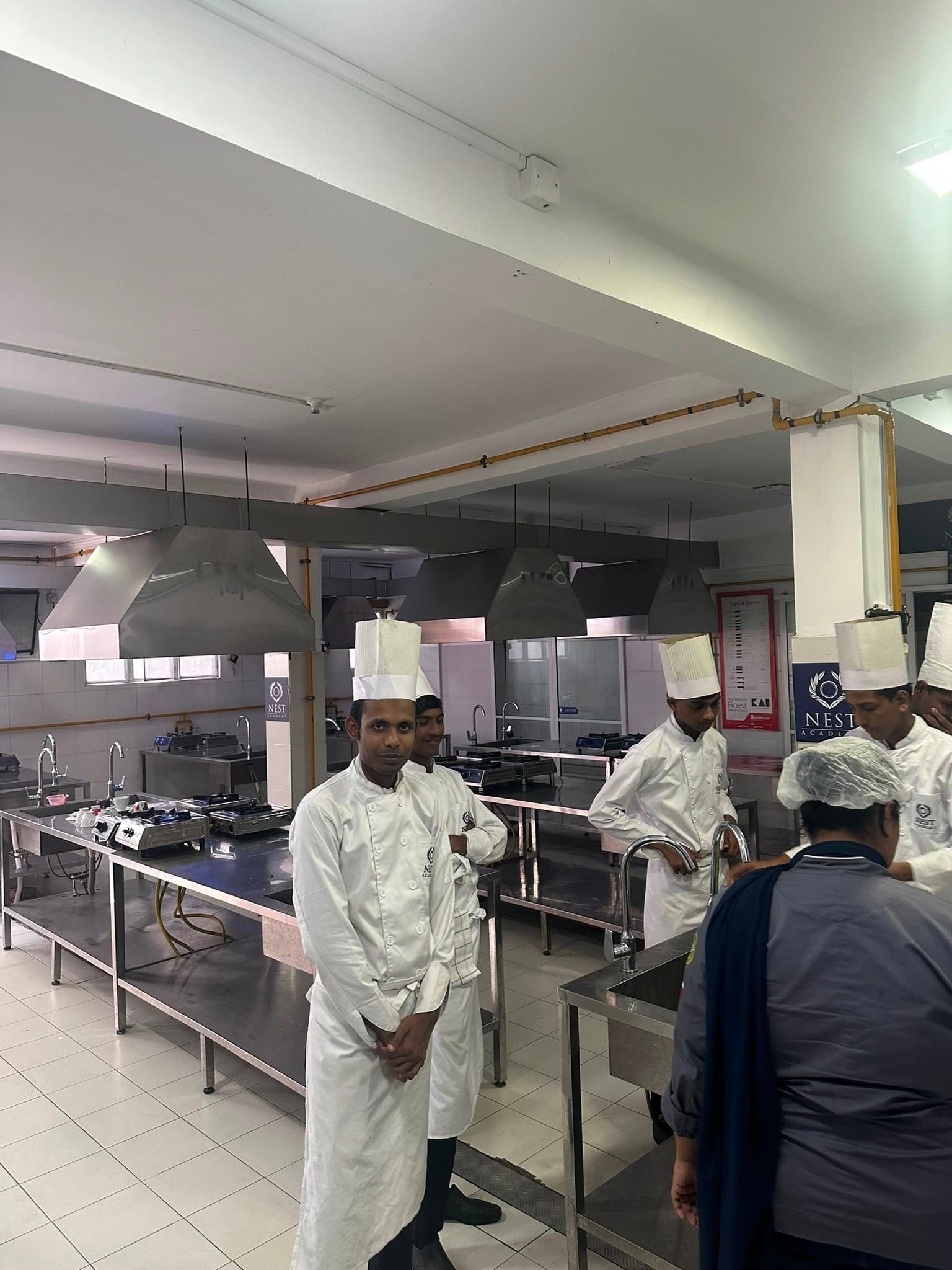 Chefs in white jackets and tall hats standing among the steel workstations of a training kitchen