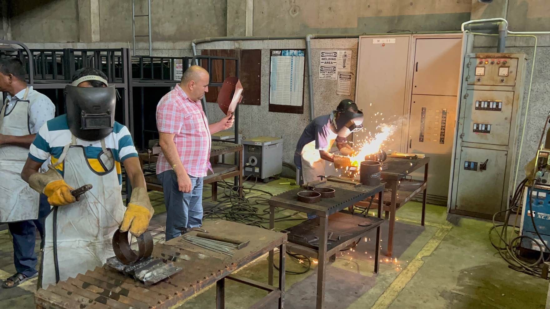 Welders in masks and leather aprons at the benches of a welding workshop, sparks flying from one, while a man in a checked shirt watches holding a hand shield