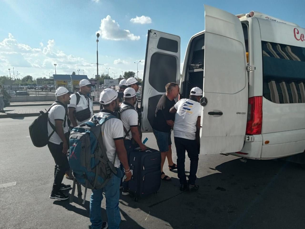 Eurozone workers in white caps, with backpacks and a suitcase, boarding a white van; one in a Eurozone Recruiting shirt stands at its open rear door.
