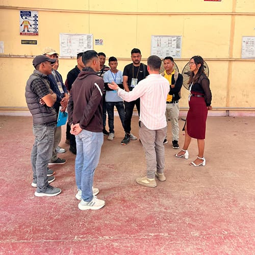 A group standing in a circle on a site yard while one man addresses them, a wall of printed safety notices behind