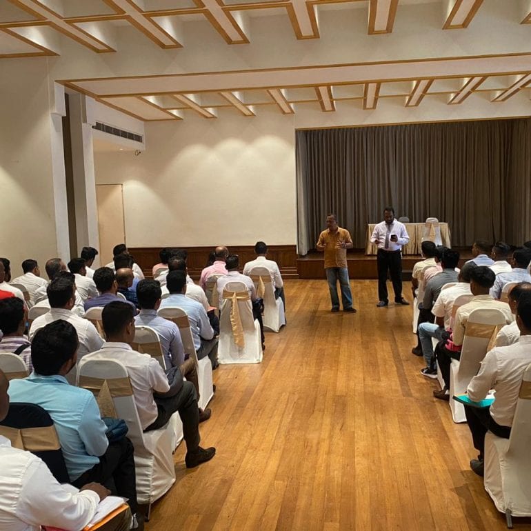 Two men addressing rows of seated candidates from the aisle of a wooden-floored function hall, a stage curtain drawn behind them