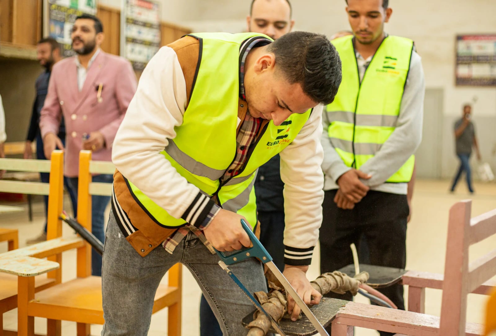 A young man in a Eurozone high-visibility vest cutting a metal bar with a hacksaw on a bench, two others in matching vests watching