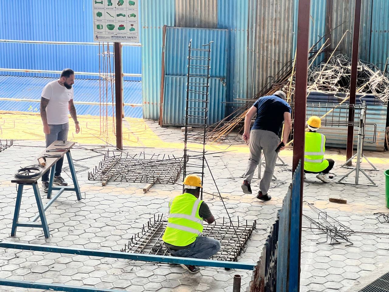 Two workers in high-visibility vests and yellow hard hats crouched over flat rebar mats on a paved yard, tying the bars, an assembled rebar cage standing behind them