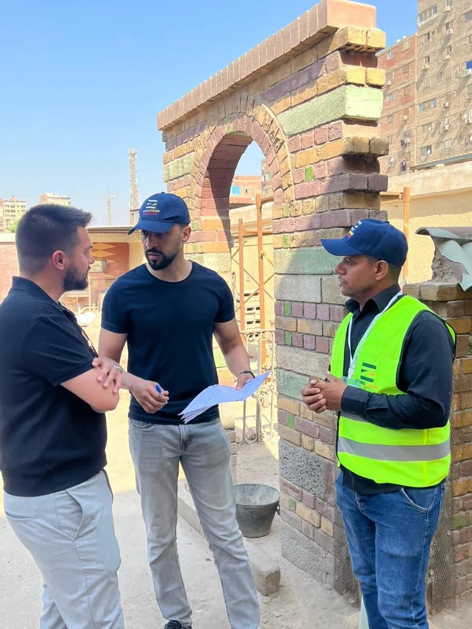 Three men talking beside a part-built brick archway on a construction site, one holding a sheaf of documents, one in a Eurozone high-visibility vest